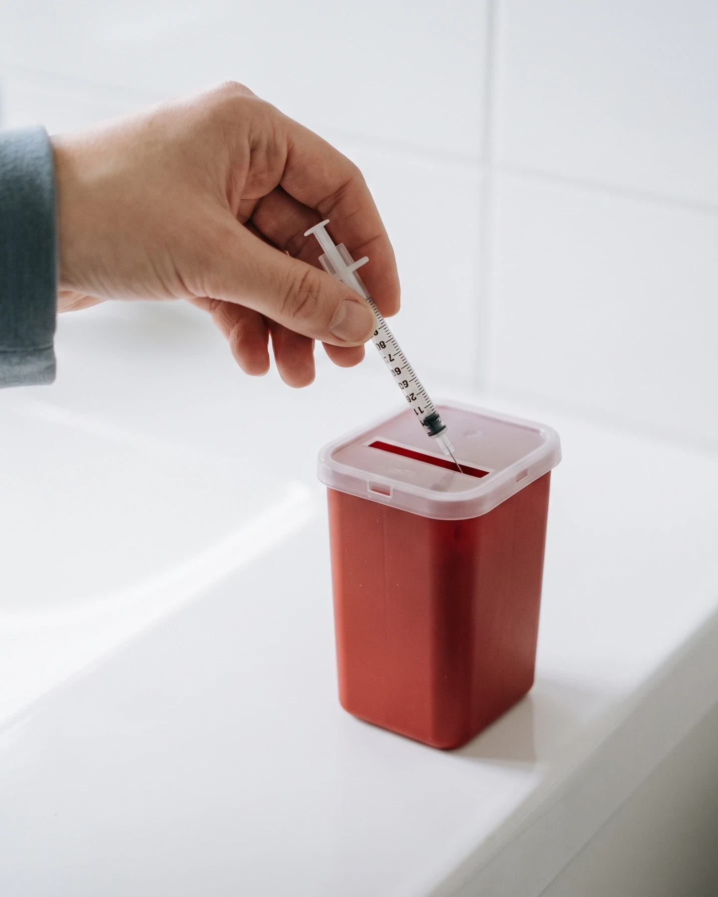 A hand dropping a used insulin syringe into a red sharps container