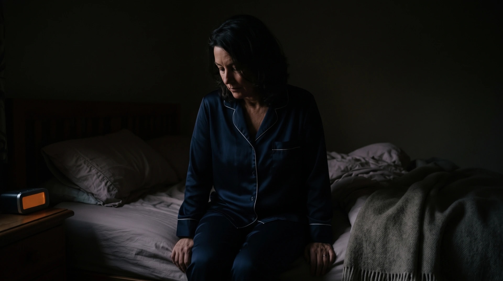 A woman sitting awake on the edge of her bed in a dark bedroom